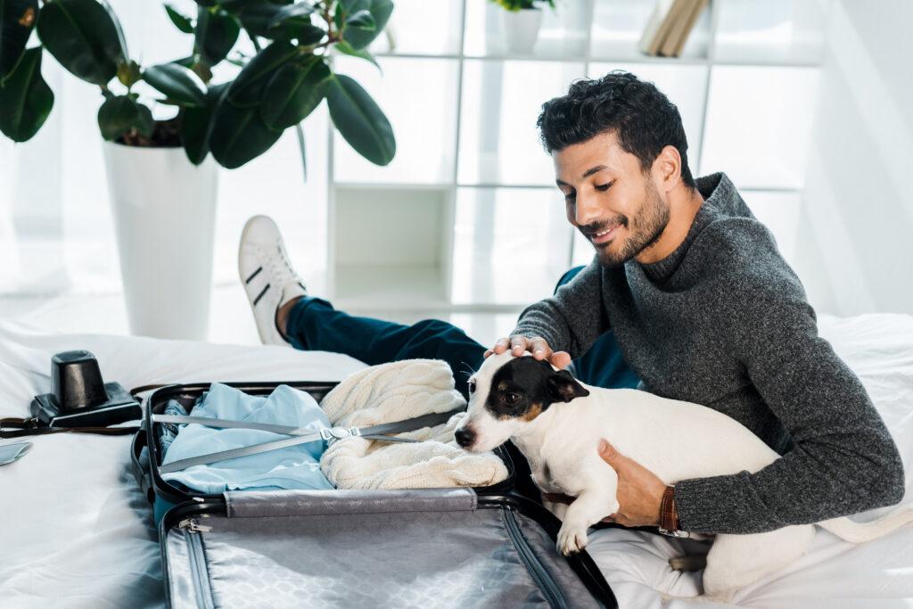 A man sitting on a bed smiles down at his Jack Russell Terrier, who is sitting in an open suitcase filled with clothes, preparing for travel.