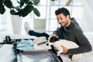 A man sitting on a bed smiles down at his Jack Russell Terrier, who is sitting in an open suitcase filled with clothes, preparing for travel.